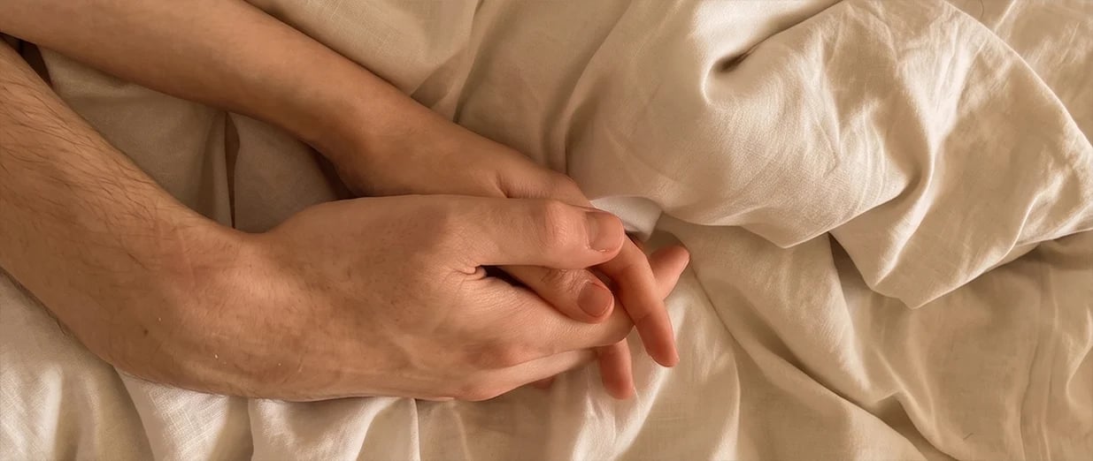 Two people holding hands on soft, light-colored bed sheets