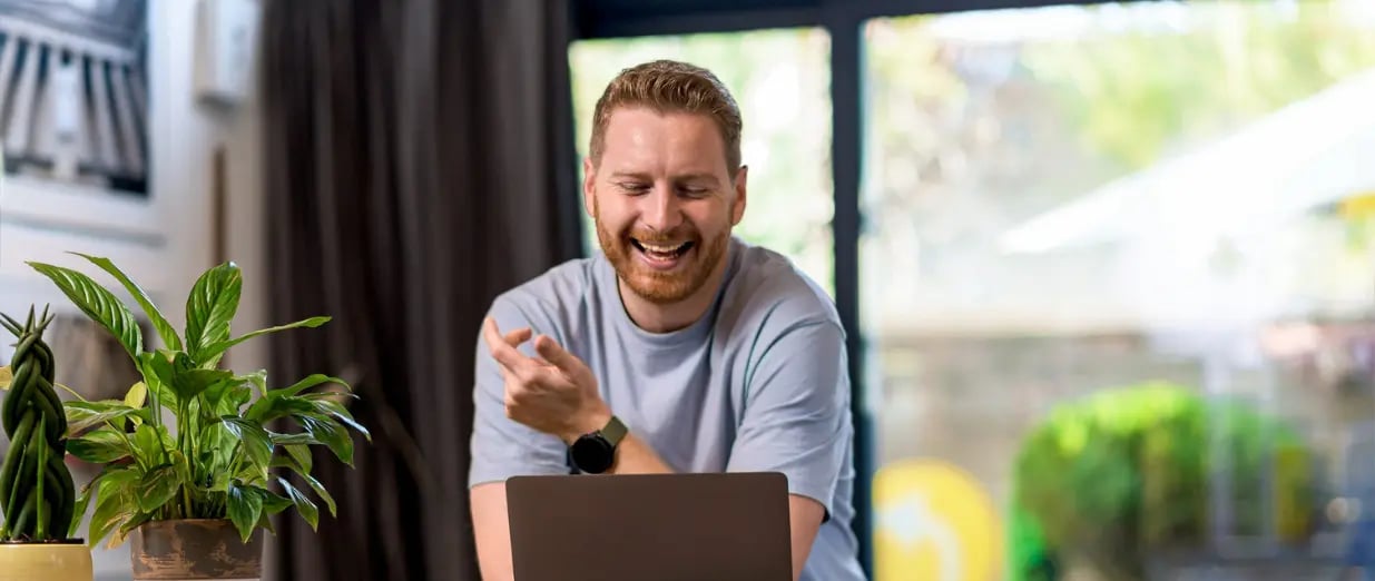 Ein lachender Mann sitzt vor einem Laptop im Wohnzimmer
