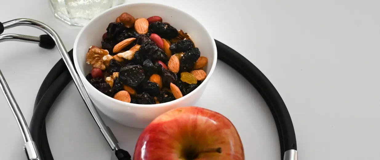 Sch&uuml;ssel mit Trockenfr&uuml;chten und N&uuml;ssen, roter Apfel und Stethoskop auf wei&szlig;em Tisch