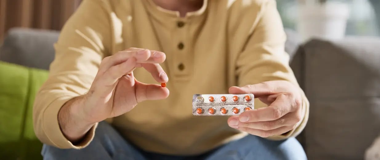 Ein Mann hält orangefarbene Tabletten in einer Blisterpackung über einem Glas Wasser auf dem Tisch
