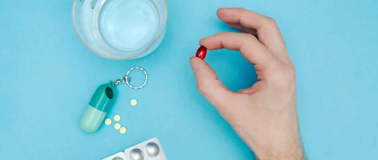 A hand holding a red capsule next to water and pills
