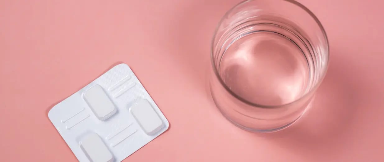 Blisterpackung mit Tabletten neben einem Glas Wasser auf rosa Hintergrund