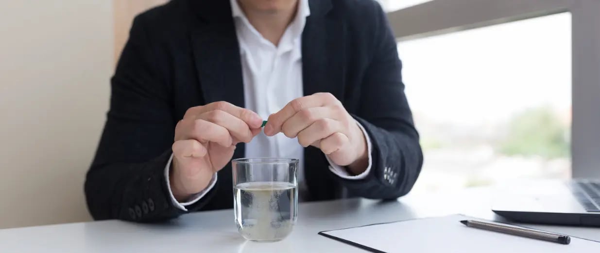 Man in business attire taking a pill with a glass of water at a desk