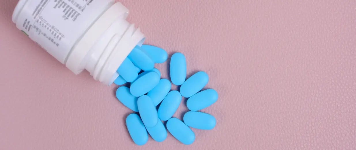 Blaue Tabletten liegen aus einer ge&ouml;ffneten Medikamentenflasche auf rosa Hintergrund
