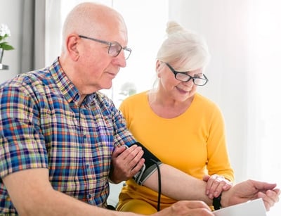 Ein älterer Mann misst seinen Blutdruck mit Hilfe seiner Frau