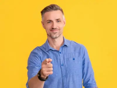 A smiling man points directly at the camera against a yellow background