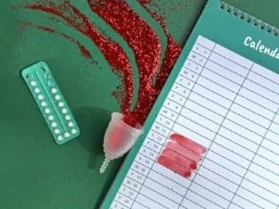 Um copo menstrual com purpurinas vermelhas, um despertador, a pílula contracetiva e um calendário com os dias menstruais marcados a vermelho