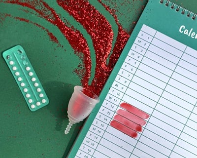 Eine Menstruationstasse mit rotem Glitzer, ein Wecker, Verhütungspillen und ein Kalender mit rot markierten Menstruationstagen