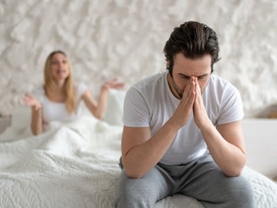 Ein Mann sitzt deprimiert auf dem Bett, während seine Frau unzufrieden ist und ihn anschreit.