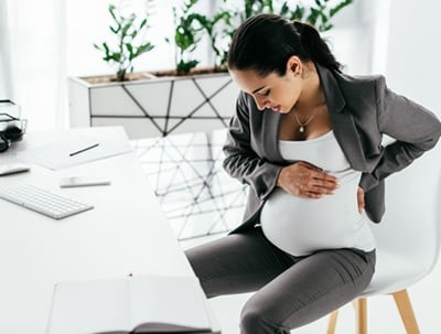 Eine schwangere Frau sitzt im Office, hält ihren Bauch mit den Händen und erträgt Schmerzen