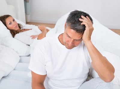 Ein Mann sitzt mit Kopfschmerzen im Bett, während seine Frau auf ihn wartet.