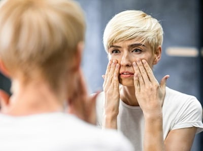  Frau betrachtet Falten und berührt die Haut unter den Augen im Badezimmer